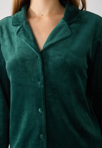 Grønn fleece pysjamasoverdel med sjalkrave, lange ermer og mykt avrundede knappelukkinger. Stoffet ser plyschaktig og glatt ut.