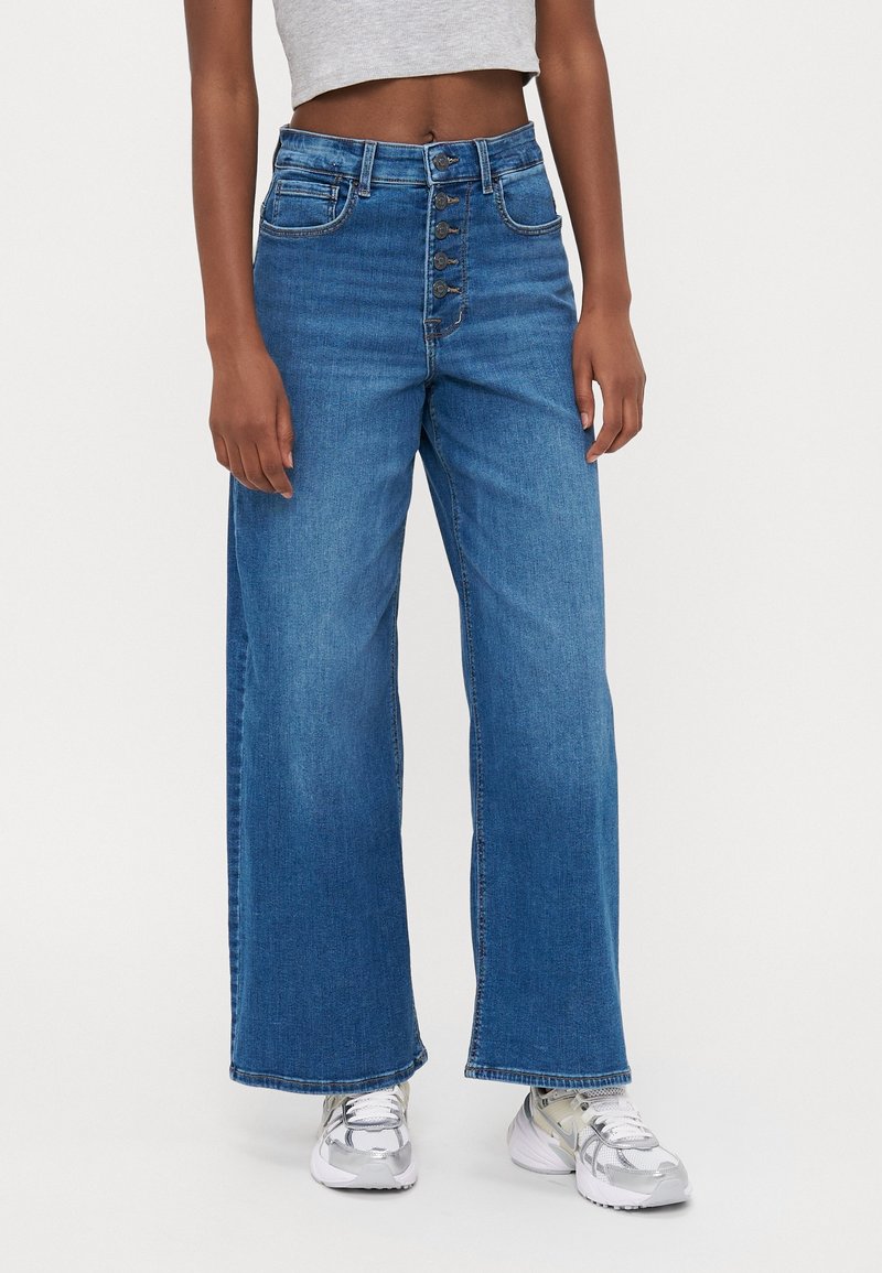 Personne portant un jean bleu taille haute à jambes larges avec cinq boutons sur le devant et des baskets argentées, debout devant un fond uni.