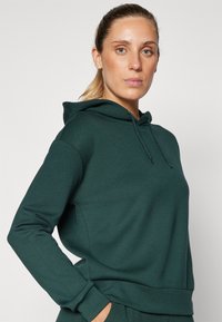 Sweatshirt à capuche en tissu vert foncé, présentant une coupe décontractée, une poche kangourou et des poignets côtelés. Texture lisse avec cordons de serrage réglables.