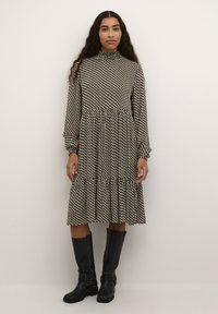 Robe avec un col haut et des manches longues, fabriquée en tissu noir et blanc à motifs, avec une jupe à volants et des poignets élastiques. Bottes noires jusqu'aux genoux.