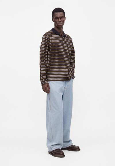 Homme debout portant un polo à manches longues rayé marron et beige, un jean bleu clair à jambes larges et des chaussures marron foncé sur fond blanc.