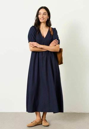 Frau mit langen dunklen Haaren, die ein marineblaues Midikleid, beige Ballerinas und eine braune Schultertasche trägt, steht mit verschränkten Armen und lächelt.