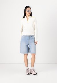 Hvit langermet zip-up topp med gule detaljer, lyseblå denimshorts, og grå joggesko med rosa detaljer. Grå solbriller.