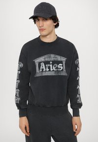 Sort sweatshirt med et stort "Aries" logo i arkitektonisk design, lange ærmer med gentagen tekst, blød tekstur og ribbede manchetter.
