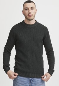 Mørkegrønn strikkegenser med rund hals, teksturert stoff, ribbestrikkede mansjetter og kant; brukt over lyseblå jeans.
