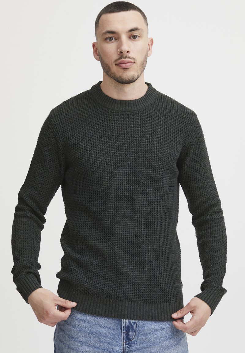 Mørkegrønn strikkegenser med rund hals, teksturert stoff, ribbestrikkede mansjetter og kant; brukt over lyseblå jeans.