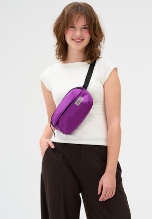 Junge Frau in weißem Oberteil und dunkler Hose mit einer violetten Umhängetasche mit schwarzem Riemen, die vor einem einfarbigen Hintergrund lächelt.