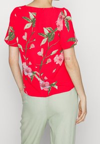 Blusa rossa con maniche corte a palloncino, caratterizzata da un motivo floreale in tonalità di rosa e verde, abbinata a pantaloni verde chiaro.