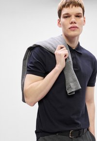 Μπλε πόλο μπλουζάκι με κοντά μανίκια, υφασμάτινη υφή, τριών κουμπιών. Ανοιχτό γκρι πουλόβερ περασμένο στους ώμους, με ριχτή μανσέτα.