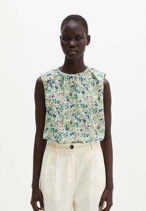 Femme portant un chemisier sans manches à fleurs bleues et vertes, associé à un pantalon crème taille haute avec une fermeture par bouton.
