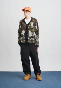 Pleteni kardigan s uzorcima u smeđoj, crnoj i bijeloj boji; duži rukavi, V-izrez, crni gumbi, uparen s crnim cargo hlačama i narančastim kapom.