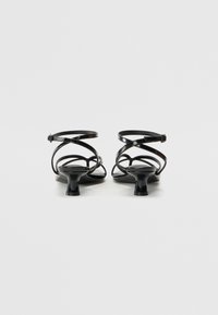 Sandales à talons en cuir verni noir avec des brides croisées autour de la cheville, bout ouvert et un petit talon carré. Texture lisse et design minimaliste.