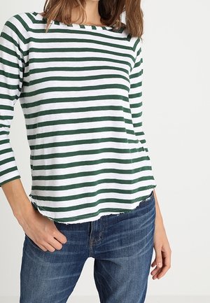 Vrouw draagt een wit-groen horizontaal gestreept shirt met lange mouwen, gecombineerd met een blauwe spijkerbroek, staand tegen een effen lichte achtergrond.