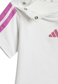 Maglietta atletica bianca con strisce rosa sulle spalle, bottoni a pressione al colletto e un logo rosa sul davanti. Tessuto in cotone con una texture liscia.