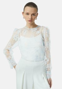 Durchsichtige weiße Bluse mit Blumenmuster und Puffärmeln, über einem schlichten weißen Tanktop getragen, kombiniert mit hochtaillierten weißen Hosen.