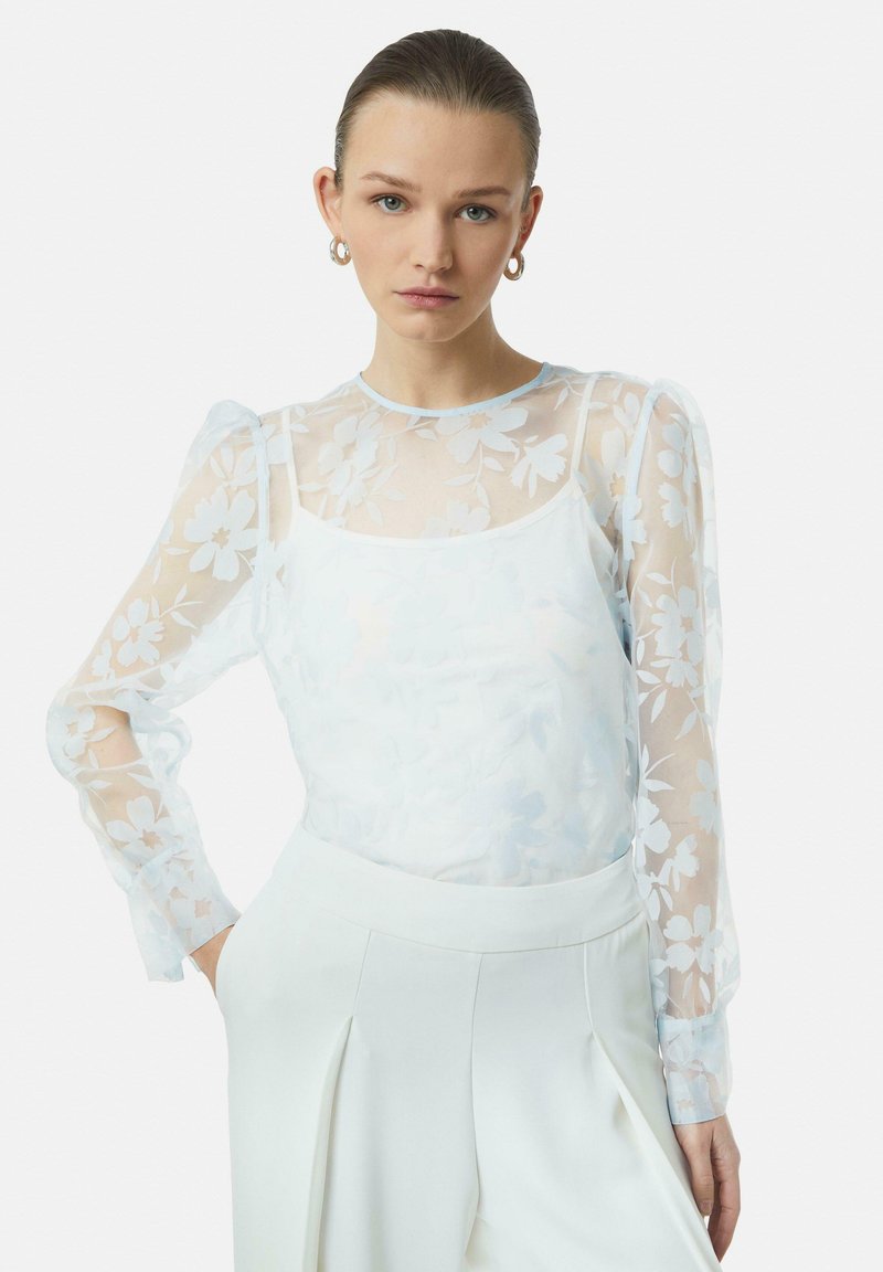 Durchsichtige weiße Bluse mit Blumenmuster und Puffärmeln, über einem schlichten weißen Tanktop getragen, kombiniert mit hochtaillierten weißen Hosen.