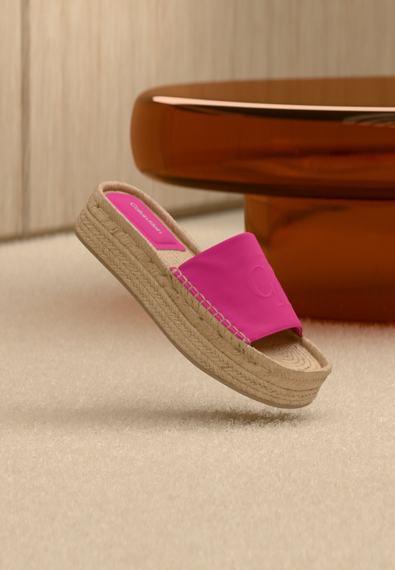 Mule espadrille rose à bout ouvert avec une semelle plateforme en jute tressé sur un tapis beige près d'une table ronde en bois.