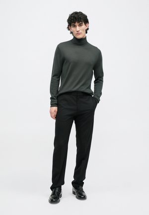 Suéter de gola alta verde escuro, ajustado, combinado com calças pretas. O modelo usa mocassins pretos. Fundo simples, cores neutras.