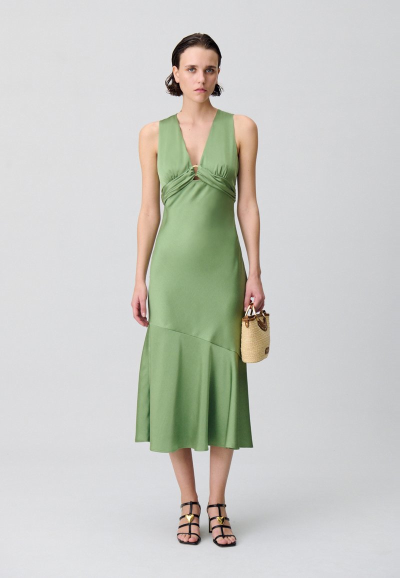Robe en satin verte avec un décolleté en V profond et un devant torsadé, ourlet évasé, portée avec des sandales noires à lanières et un petit sac tissé.