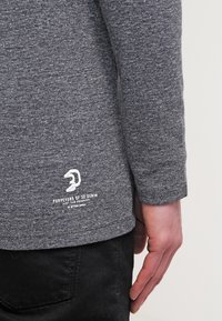 Harmaa pitkähihais shirt, jossa on tekstuuri. Oikeassa alareunassa valkoinen logo ja teksti, tumman mustissa housuissa.