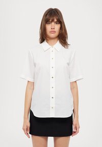 Camicia bianca a maniche corte con colletto e bottoni dorati, caratterizzata da un tessuto liscio e un orlo arrotondato.