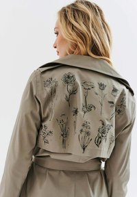 Khaki trencz z kwiatowymi ilustracjami w czarnym tuszu na plecach, z paskiem w talii i szerokim kołnierzem, wykonany z lekkiego materiału.