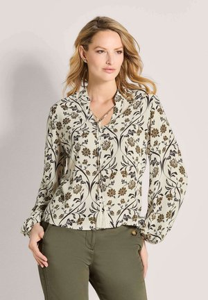 Vrouw met golvend blond haar, die een witte blouse met lange mouwen en een bruin bloemenpatroon en olijfgroene broek draagt, staand met handen in de zakken.