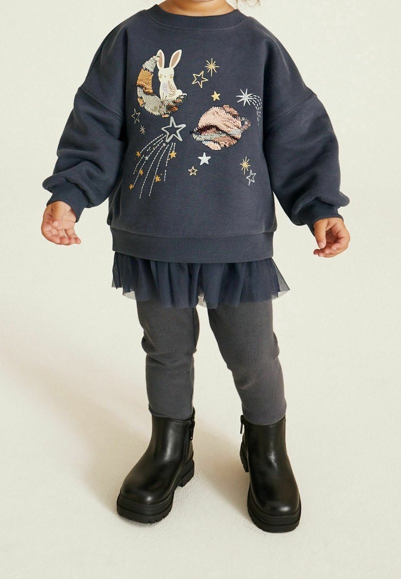 Enfant portant un sweat-shirt foncé avec un motif lapin et planètes, des leggings foncés, une jupe en tulle et des bottines noires, debout sur un sol clair.