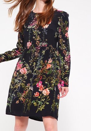 Vrouw draagt een zwarte jurk tot op de knie met lange mouwen en een kleurrijke bloemenprint met roze, rode en groene bloemen en bladeren.