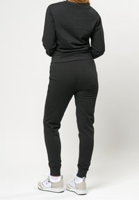 En person som bär en svart träningsdräkt och vita sneakers står med ryggen mot en enfärgad bakgrund. Deras långa, vågiga hår faller ner.