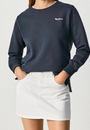Kvinde iført en mørkeblå langærmet sweatshirt med "Pepe Jeans"-logo og en hvid denimskørt, hænderne stukket ned i skørtets lommer.