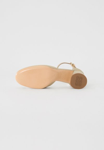 Beige Wildlederschuh mit hohen Absätzen, glänzender hellbrauner Sohle, Knöchelriemen und eingeprägtem Logo-Detail an der Unterseite.