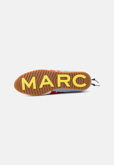 Semelle de chaussure avec une bande de roulement rainurée marron et de grandes lettres jaunes formant "MARC" sur la semelle, lacets visibles à l'arrière du talon.