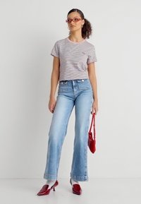 Γυναίκα που φοράει ριγέ μπλούζα, τζιν παντελόνι ψηλής μέσης σε ανοιχτό μπλε χρώμα, κόκκινα γυαλιά και τακούνια, κρατώντας μια μικρή κόκκινη τσάντα, στέκεται μπροστά σε έναν λευκό τοίχο.