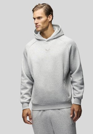 Homme portant un sweat à capuche gris clair et un pantalon de survêtement assorti, regardant vers la gauche sur un fond gris uni.
