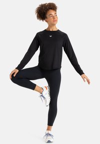 Musta pitkähihainen urheilupaita, joka on yhdistetty mustiin legginsseihin. Malli on pukeutunut valkoisiin lenkkareihin, joissa on oranssia sävyä, pitäen tasapainoista asentoa.