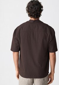 Camisa marrón de manga corta con textura y un patrón sutil, cuello y dobladillo recto. La tela parece suave y ligeramente estructurada.