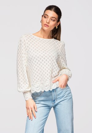 Mujer con blusa blanca de encaje de manga larga y jeans azul claro, con la mano en el bolsillo, mirando hacia su izquierda contra un fondo liso.