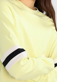 Light yellow sweatshirt featuring long sleeves with black and white striped accents at the cuffs. Soft cotton material; casual design.