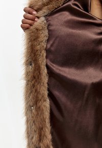 Brun satinbeklædning med en faux fur krave. Fåren har en blød tekstur, mens satinoverfladen fremstår glat og skinnende. Krystal detaljer er synlige.
