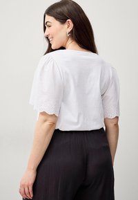 Top bianco con maniche a pompom e dettagli in pizzo traforato scallopati ai polsini, abbinato a pantaloni pieghettati scuri.