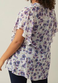 Kvinna som bär en vit blus med lila och rosa blommönster samt fjädrande ärmar, kombinerat med mörka byxor, sedd bakifrån.