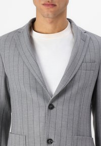 Blazer grigio a righe con colletto a revers, chiusura a un bottone e taschino sul petto a sinistra. Il tessuto ha una consistenza liscia con righe verticali.