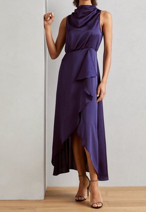 Mouwloze donkerpaarse high-low jurk met gedrapeerde halslijn en gerimpelde asymmetrische rok, gecombineerd met hoge sandalen met bandjes op een houten vloer.