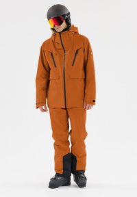 Orangene wasserdichte Skijacke mit großen Fronttaschen, Reißverschlüssen, verstellbarem Kapuze und passenden orangen Skihosen mit schwarzen Akzenten. Inklusive Skischuhe.