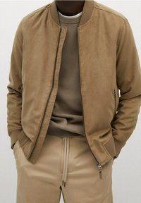 Brun ruskinds bomberjakke med lynlås foran, ribbet krave og sidelommer, parret med en struktureret beige sweater og matchende bukser.