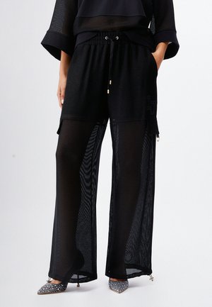 Donna con pantaloni neri trasparenti a gamba larga con tasche cargo e tacchi scintillanti a punta, mani nelle tasche.