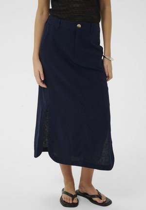 Vrouw draagt een marineblauwe midi rok met een zijsplit, een zwarte transparante top, een zilveren armband en zwarte sandalen met vierkante neus tegen een effen achtergrond.