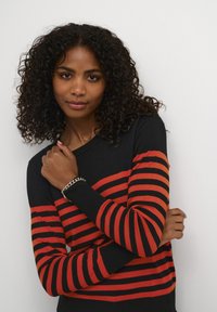 Pull à rayures noir et rouge avec des manches longues, un col rond et une texture douce, présentant un look en couches sur des bras croisés.