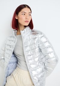 Giacca puffer argento con colletto alto, motivo trapuntato e chiusura con zip, abbinata a un top grigio chiaro e pantaloni crema.